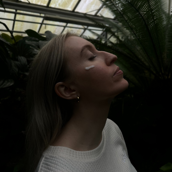 Woman applying Yui moisturizer to her cheek in a serene botanical setting, emphasizing hydration and skin nourishment.